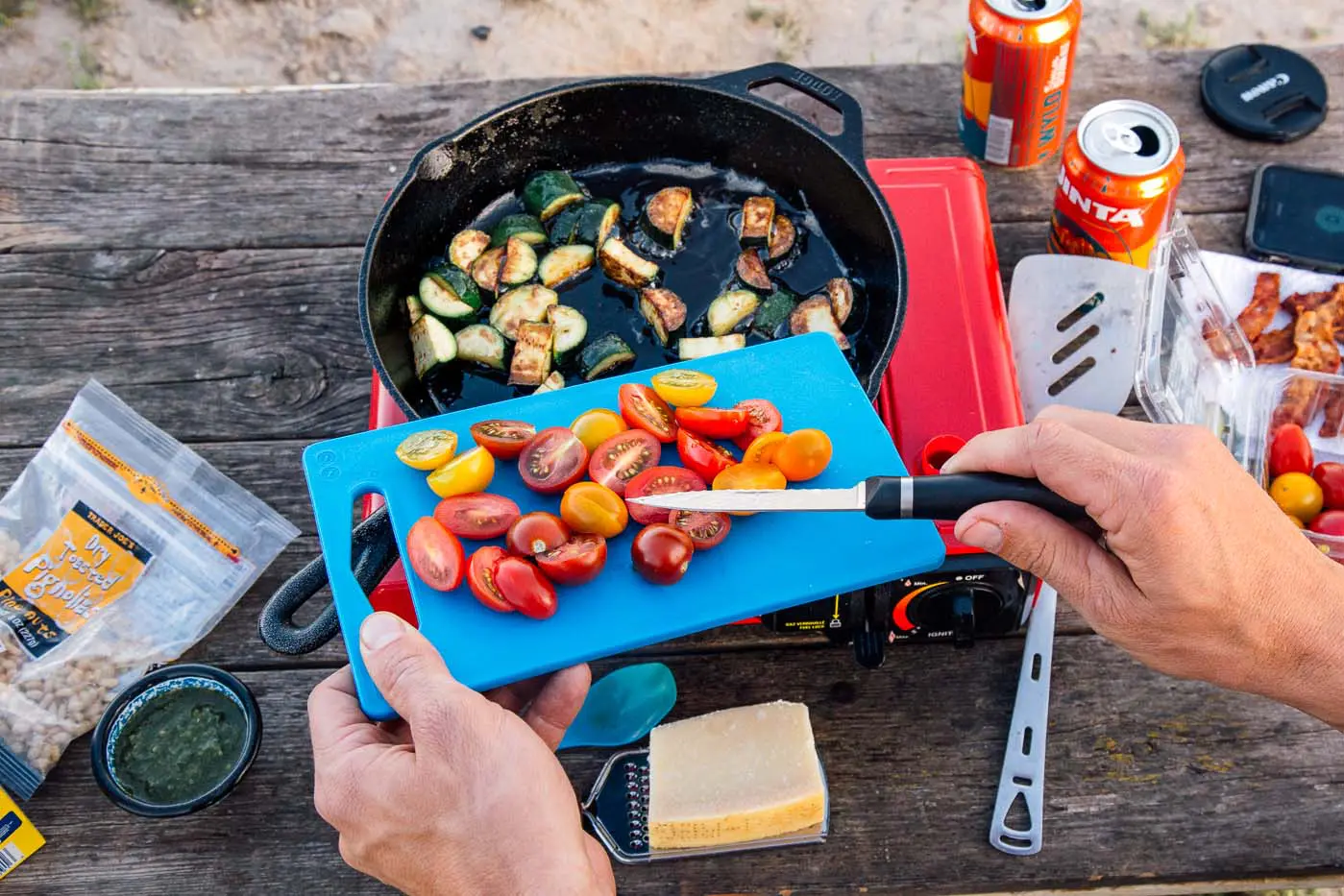 Man moving cut tomatoes from a cutting board into a cast iron skillet on a camping stove.