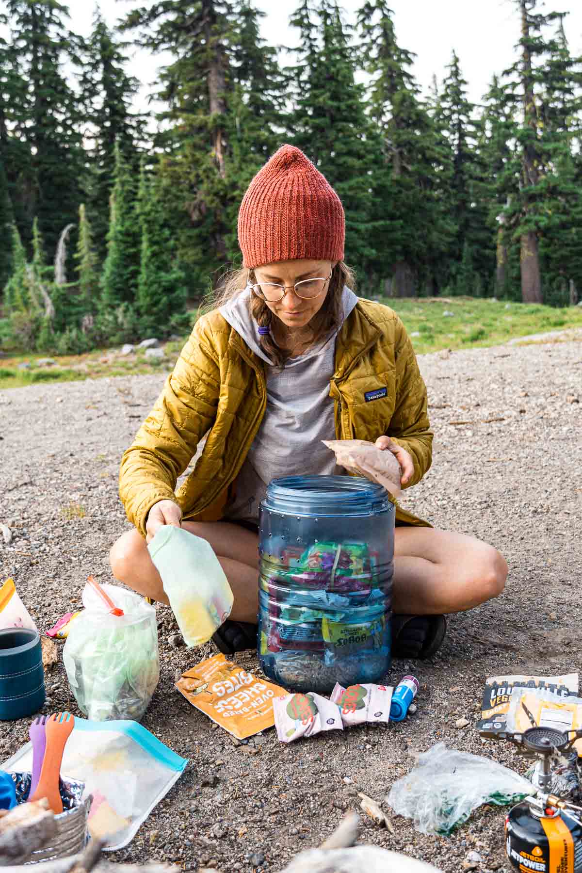 Megan is sitting on the ground with a bear canister in front of her. She is removing a backpacking meal from the canister.