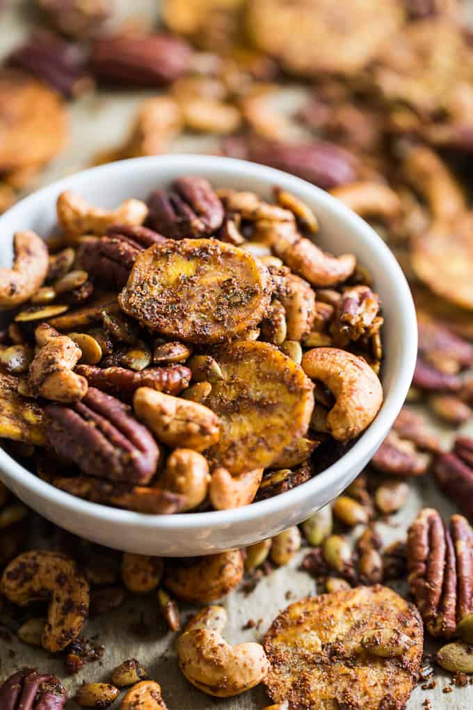 A bowl contains mixed nuts, namely pecans, cashews, and banana chips, placed amidst scattered nuts on a rustic surface.