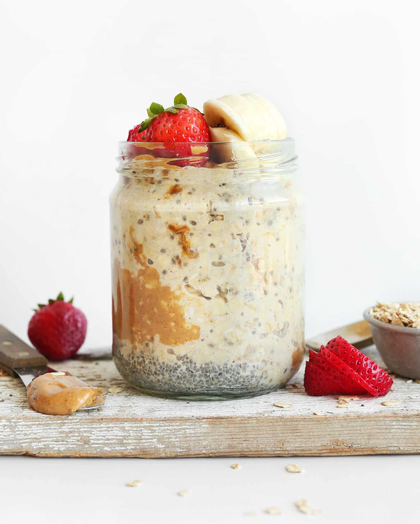 A jar filled with layers of overnight oats, topped with fresh strawberries and banana slices, sits on a rustic wooden table surrounded by scattered ingredients.