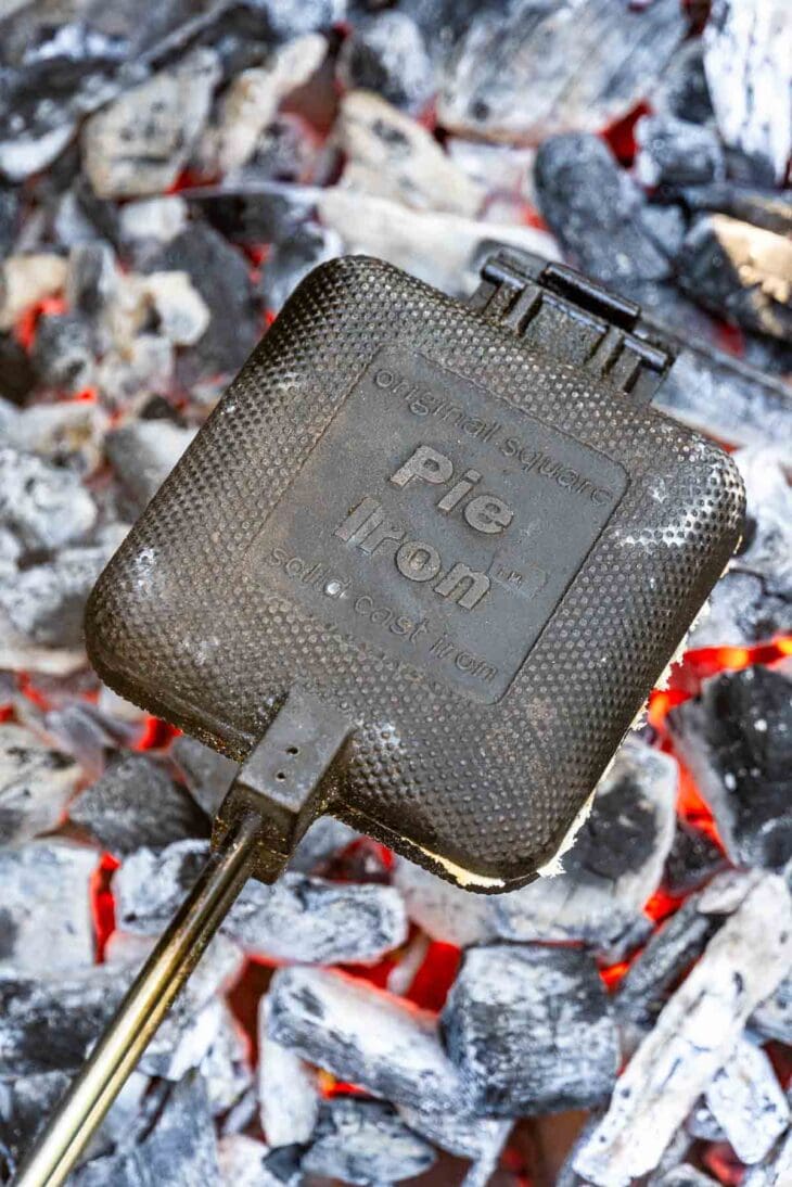 A square cast iron pie iron with a long handle sits on hot charcoal embers, showing its textured surface and brand label.