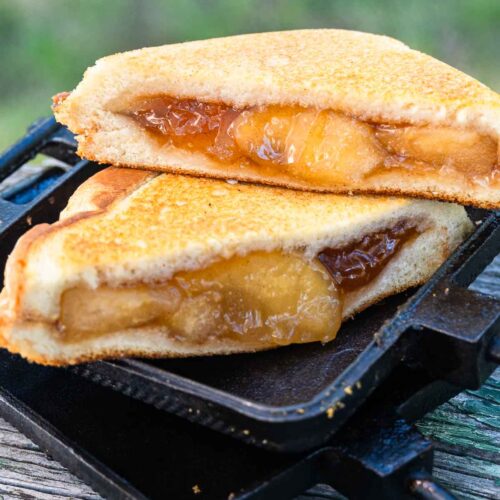 Two halves of a toasted sandwich with golden brown crust and gooey apple filling sit on a cast iron press on a wooden surface outdoors.