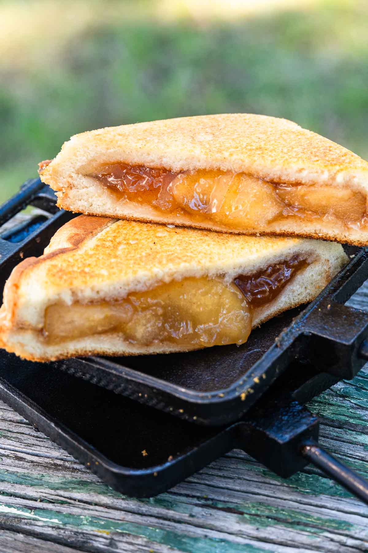 Two halves of a toasted sandwich with golden brown crust and gooey apple filling sit on a cast iron press on a wooden surface outdoors.