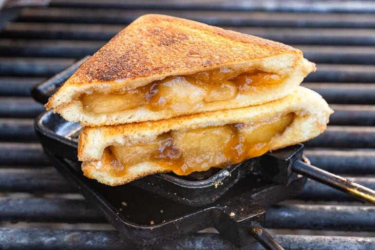 Two slices of golden-brown toasted bread with baked apple filling are stacked on a metal grill, edges crisp and caramelized.