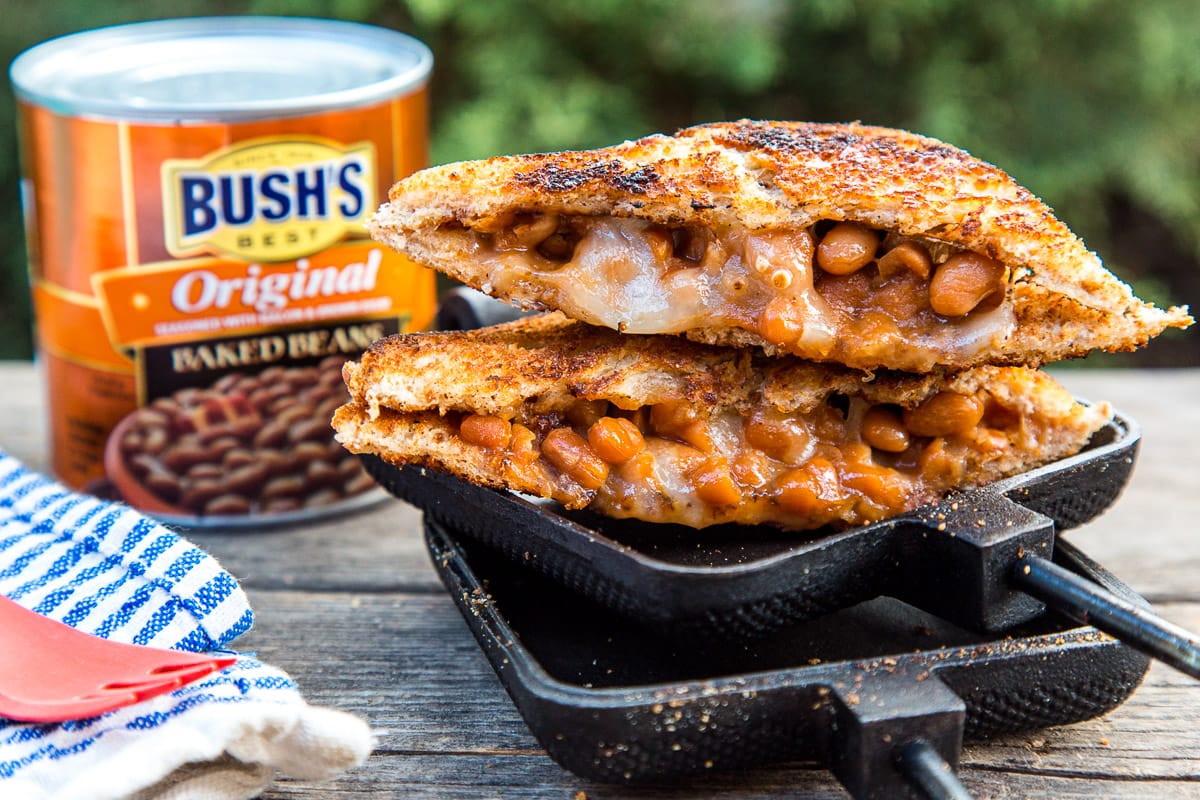 A sandwich filled with beans and cheese cut diagonally in half, resting on an open pie iron with a can of BUSH'S® Baked Beans in the background