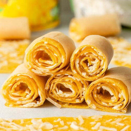 Five rolled mango fruit leathers filled with shredded coconut are stacked in a pyramid on a white surface, with extra rolls and shredded coconut around them and a blurred can in the background.