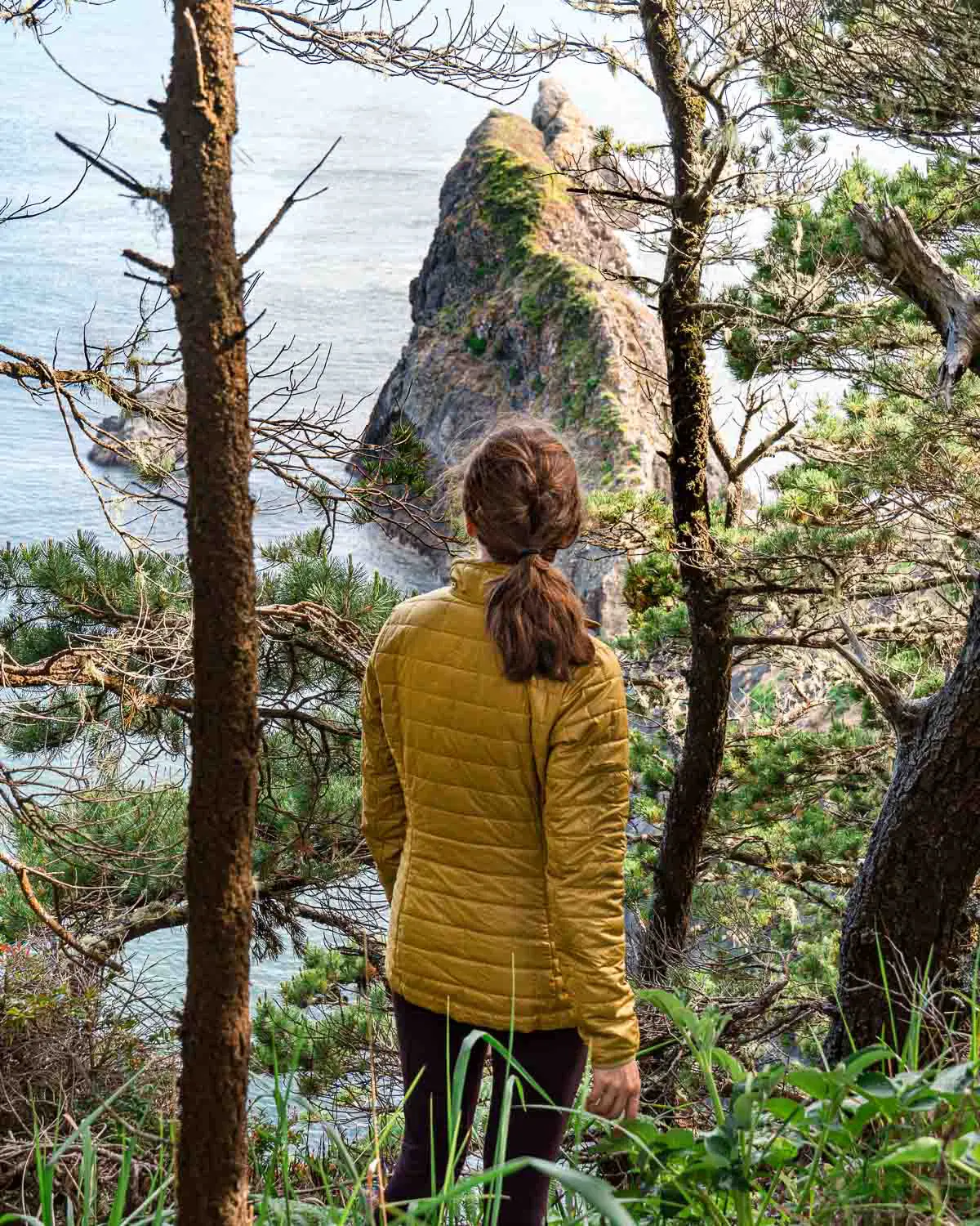 Megan stands among trees and looks out at a rock formation rising from the ocean
