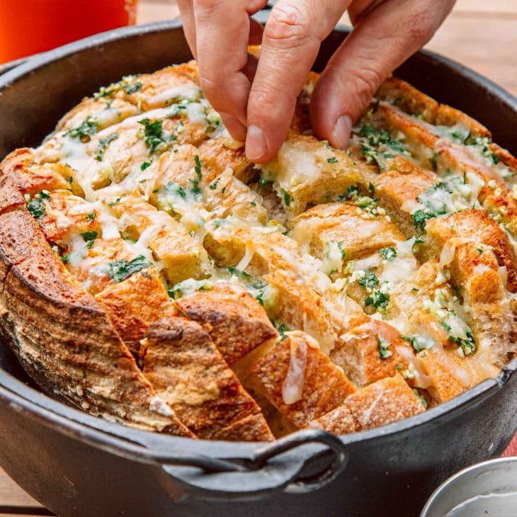 Pull apart garlic bread in a Dutch oven. a hand is reaching in to pick up a piece