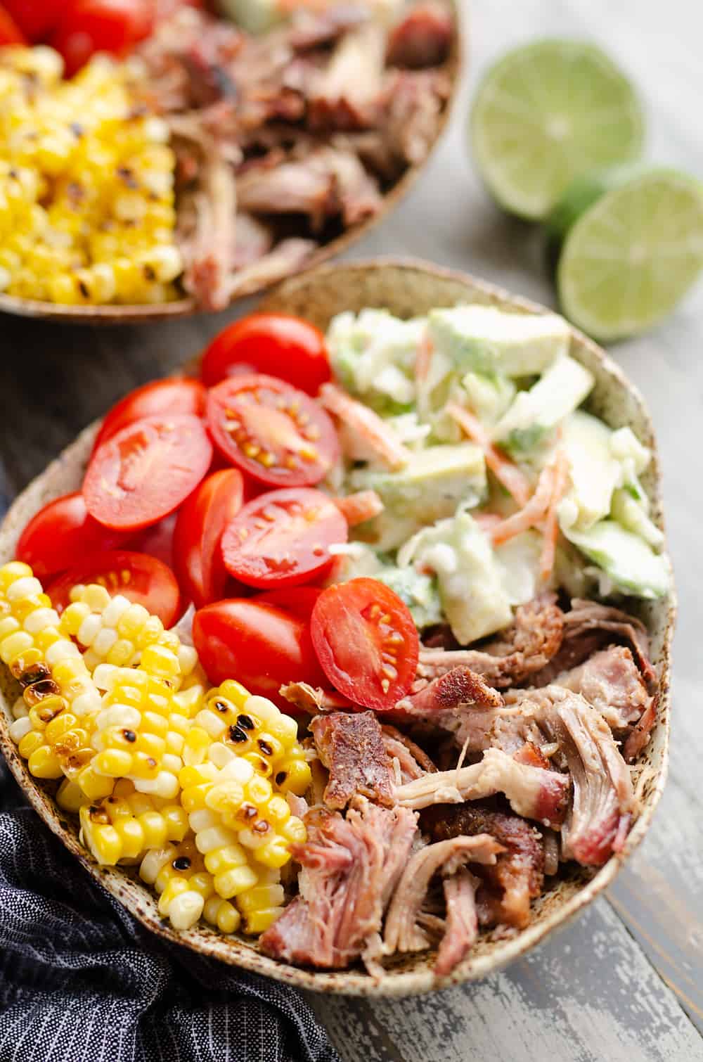 A vibrant burrito bowl on a rustic wooden surface, containing pulled pork, sliced cherry tomatoes, corn kernels, creamy coleslaw and garnished with lime wedges.