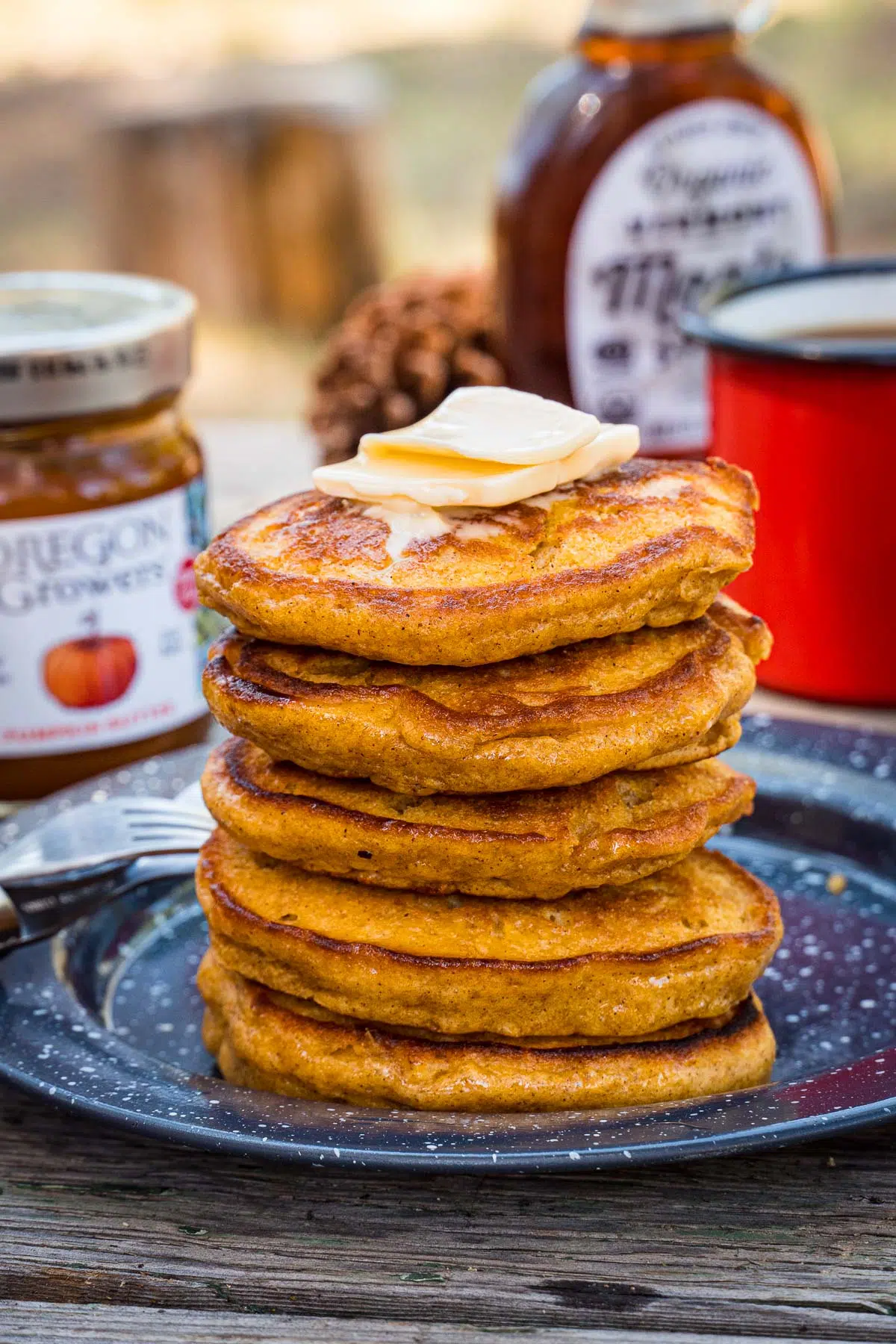 Pumpkin pancakes stacked on a plate. A coffee cup and a bottle of maple syrup are in the background.