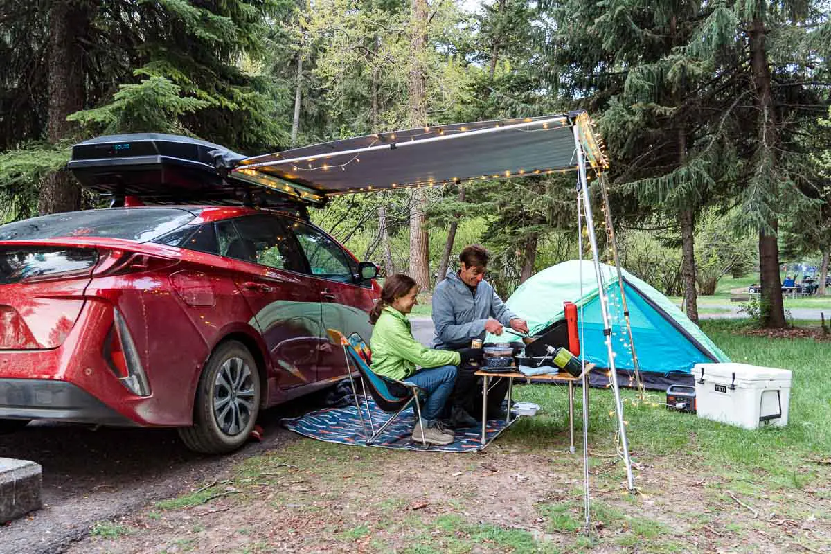 Megan and Michael sit next to a red car and are cooking on a camp stove.