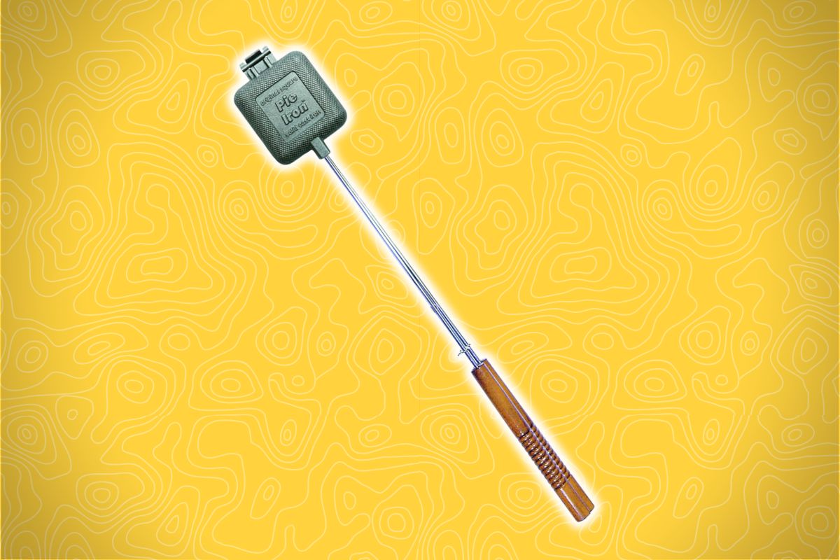 A metal pie iron with a wooden handle sits on a yellow background with swirl patterns; "Pie Iron" is engraved on its square surface.