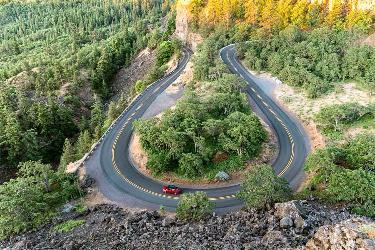 A red car driving on a U shaped road lined with trees.