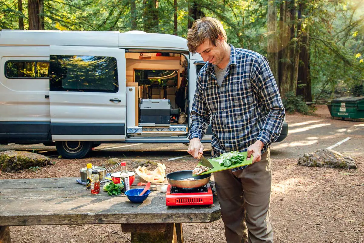 Michael adding vegetables to a skillet with a campervan and trees in the background.