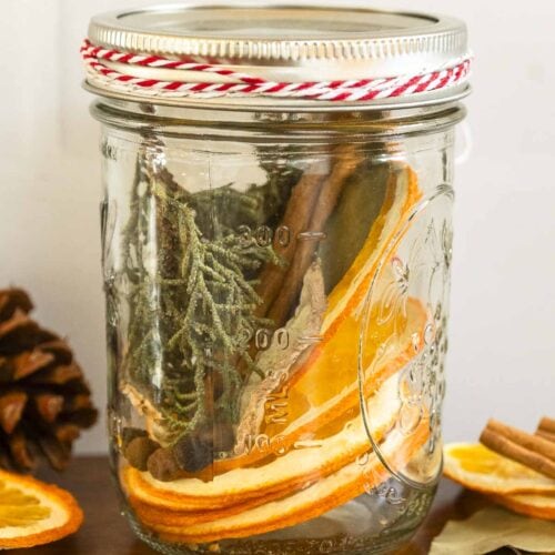 A mason jar with dried orange slices, cinnamon sticks, and greenery sits on wood beside a pinecone and extra dried orange slices.