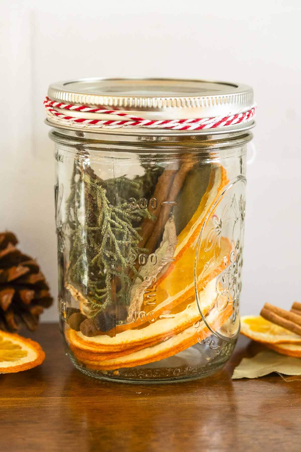 A mason jar with dried orange slices, cinnamon sticks, and greenery sits on wood beside a pinecone and extra dried orange slices.