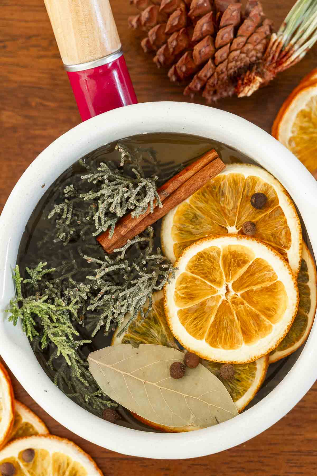 A white pot with a red handle holds water, orange slices, spices, bay leaf, and evergreen sprigs on wood, with decor nearby.