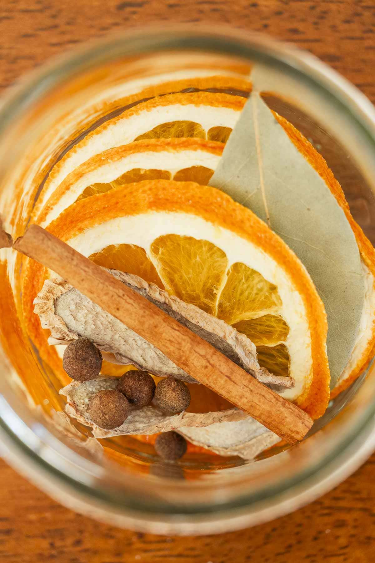 Dried orange slices, bay leaf, cinnamon stick, whole allspice, and dried ginger pieces in a glass jar on a wooden surface.