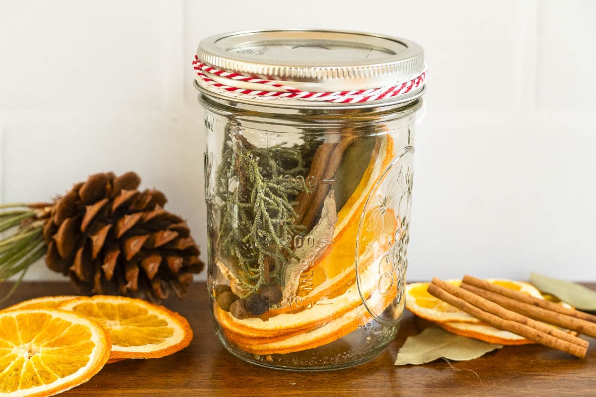 A mason jar with dried orange slices, cinnamon sticks, and greenery sits on wood, tied with red and white string; items scattered nearby.