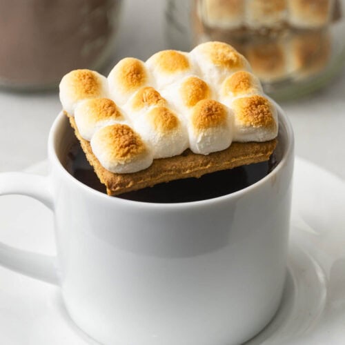 A white mug of hot chocolate is topped with a graham cracker and toasted mini marshmallows, with blurred jars in the background.