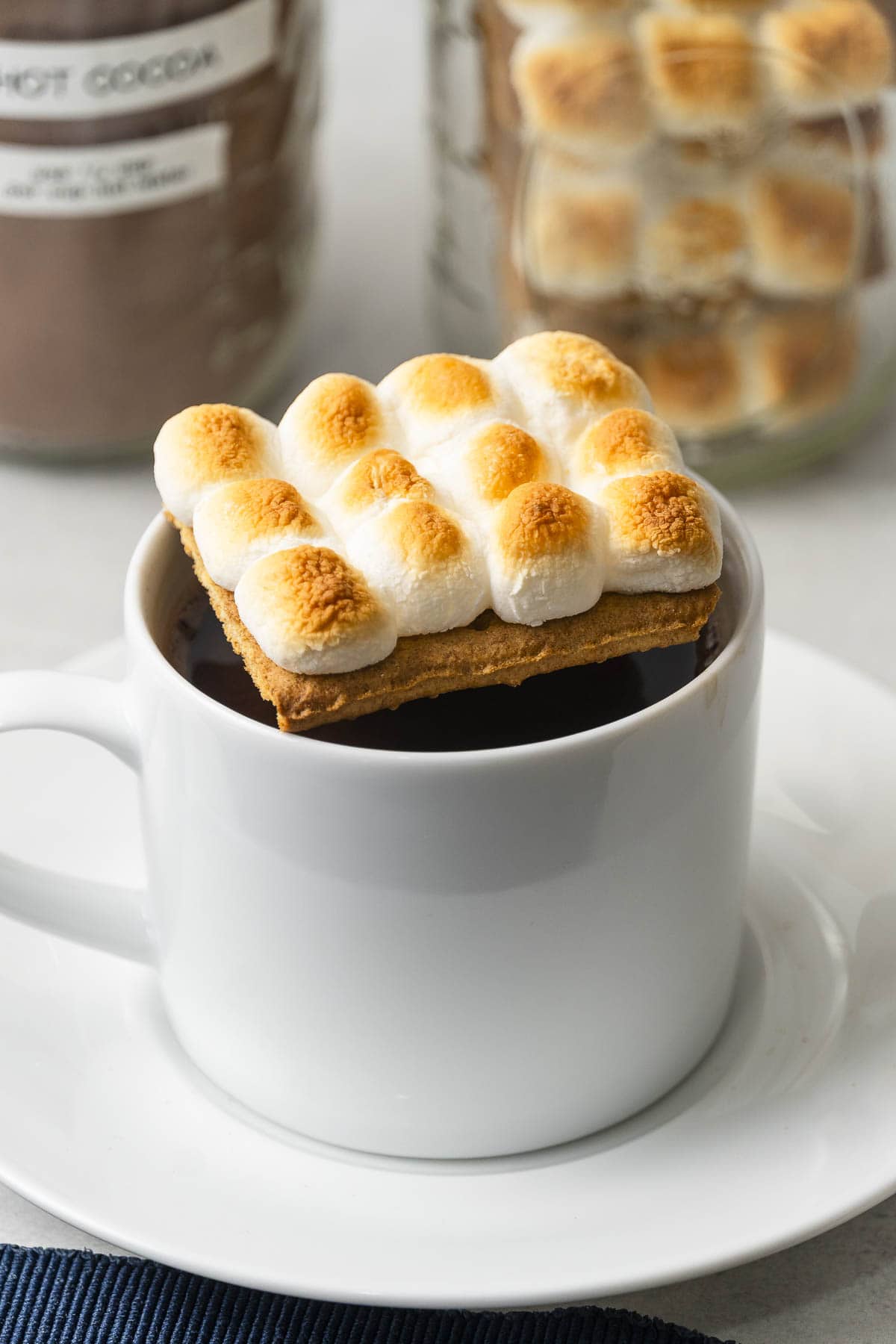 A white mug of hot chocolate is topped with a graham cracker and toasted mini marshmallows, with blurred jars in the background.
