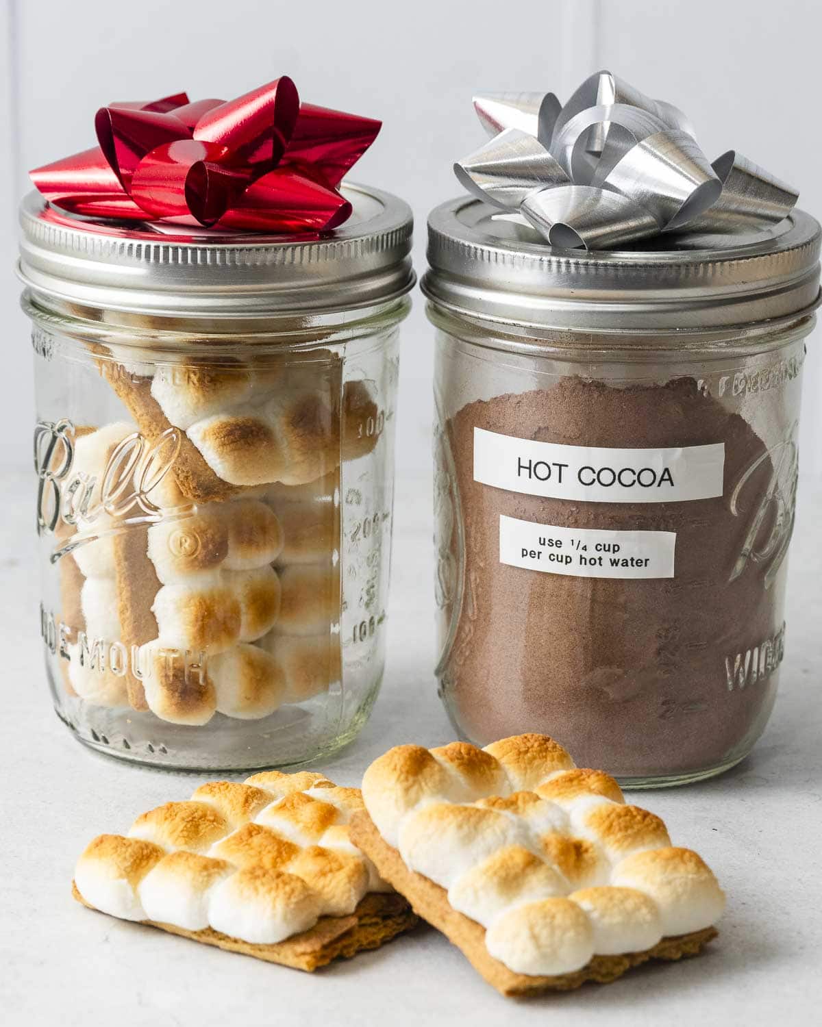 Two glass jars with metallic lids and bows; one holds s’mores, the other labeled "HOT COCOA," with two s’mores in front.