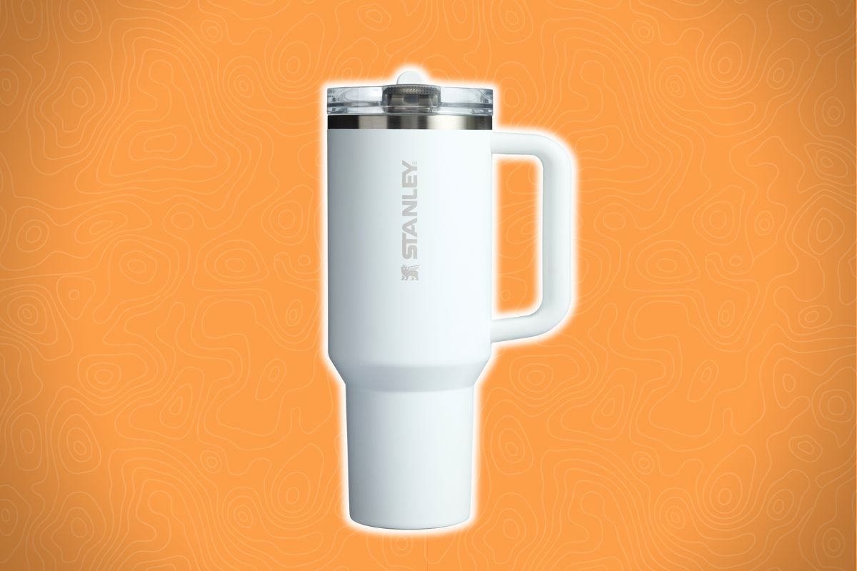 A white Stanley travel mug with a handle and clear lid sits in front of an orange background with a wavy line pattern.