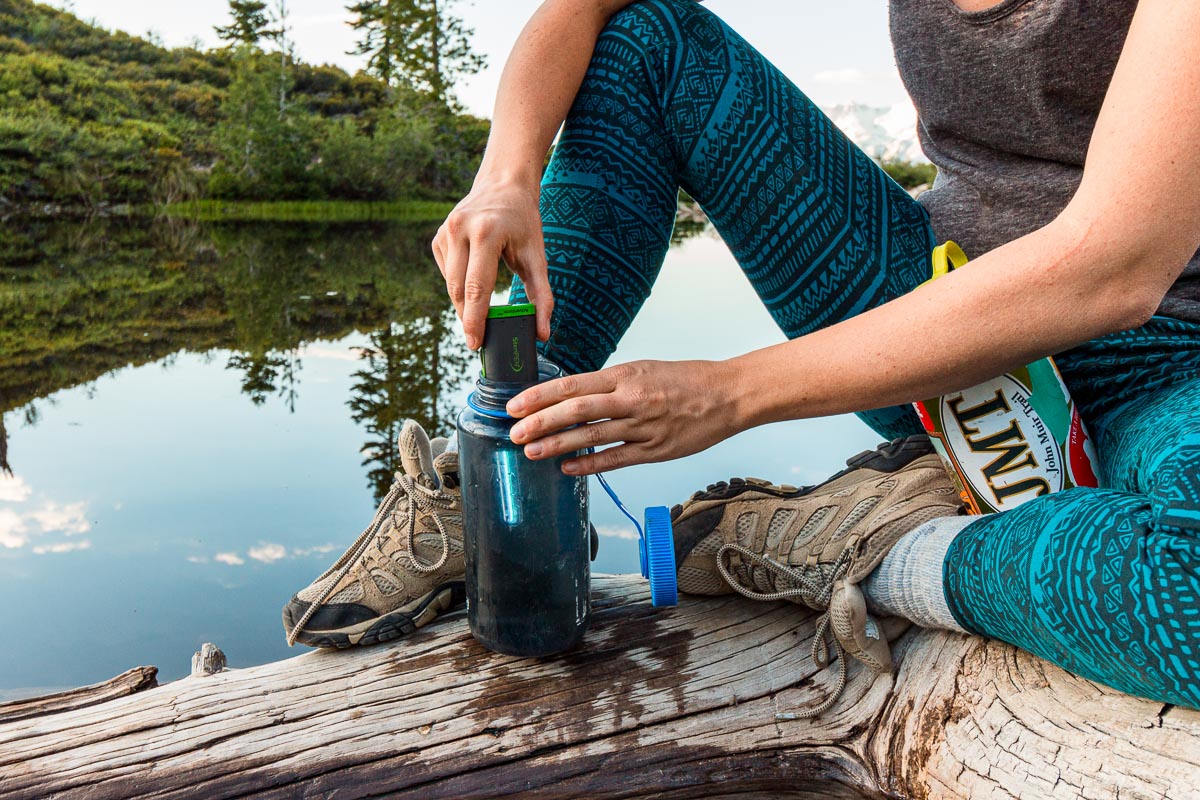Megan is sitting on a log by a lake. She is using a Steripen in a bottle of water.