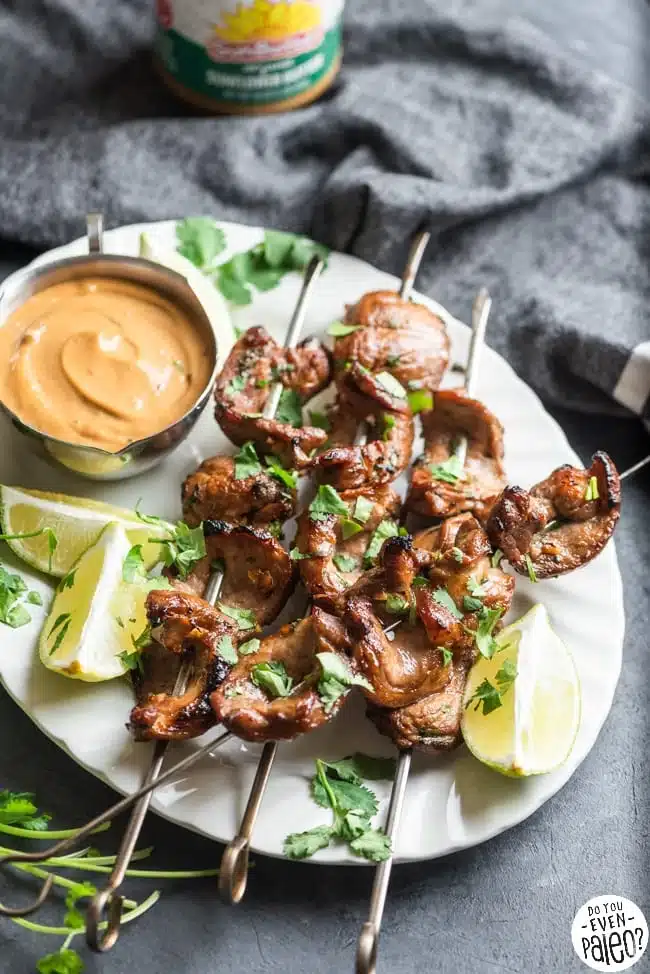 Grilled skewered meats garnished with fresh herbs and lime wedges, served with a creamy dipping sauce.