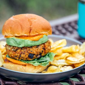A sweet potato black bean burger stacked between two buns, avocado, and spicy mayo on a plate with a natural backdrop.