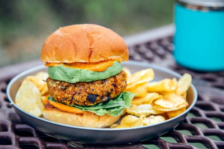 A sweet potato black bean burger stacked between two buns, avocado, and spicy mayo on a plate with a natural backdrop.