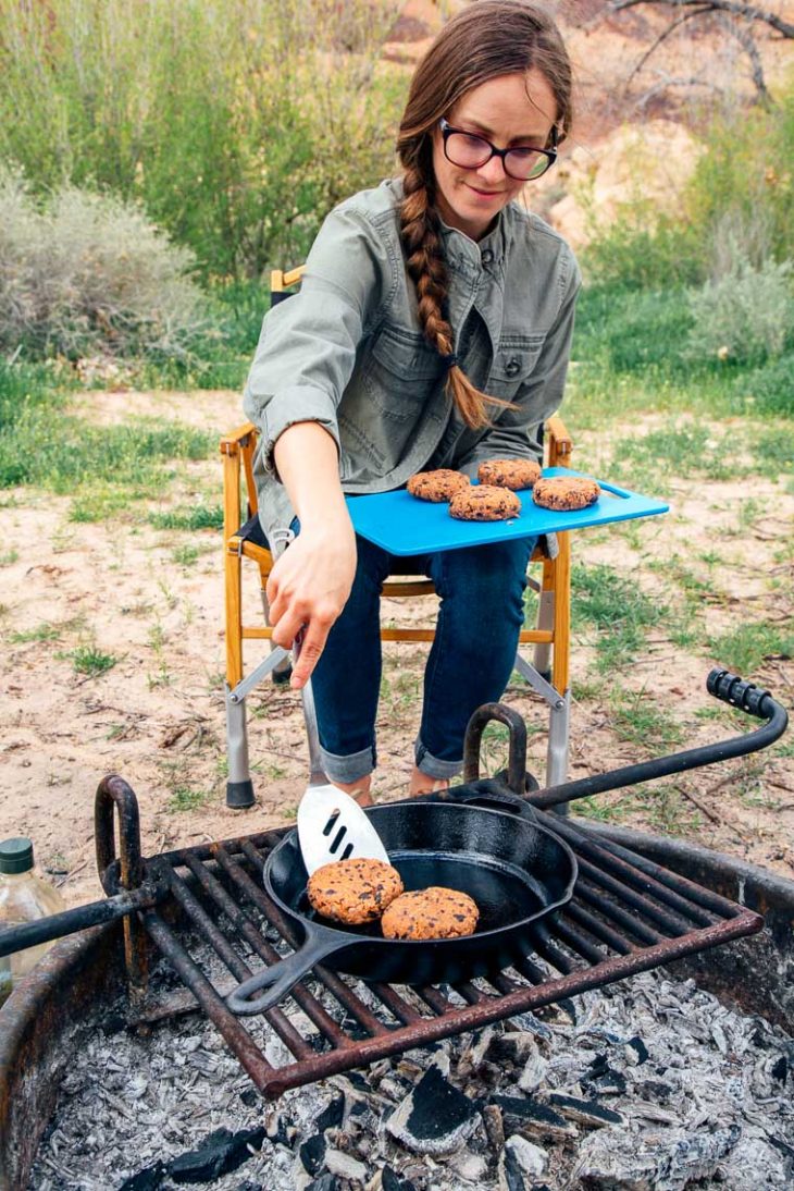 Megan is transferring black bean burger patties from a cutting board into a skillet over the campfire.
