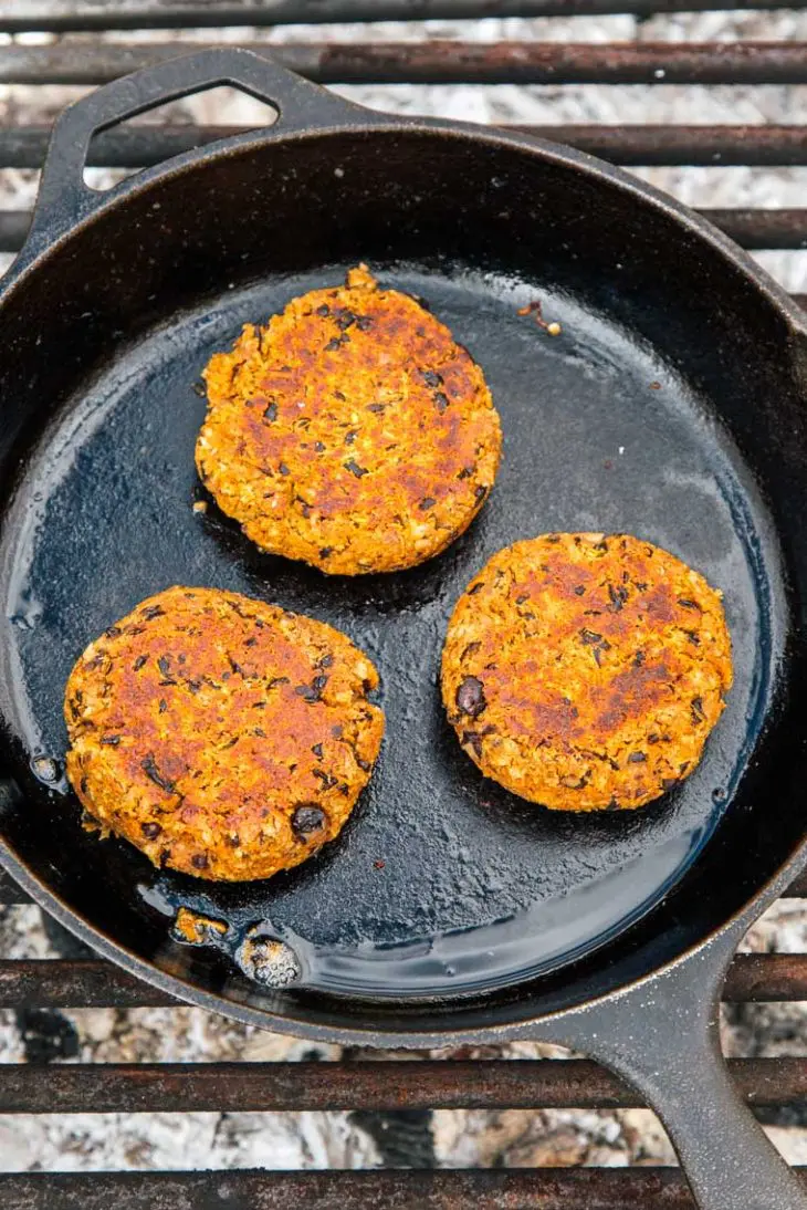 Three black bean burger patties cooking in a cast iron skillet over a campfire.