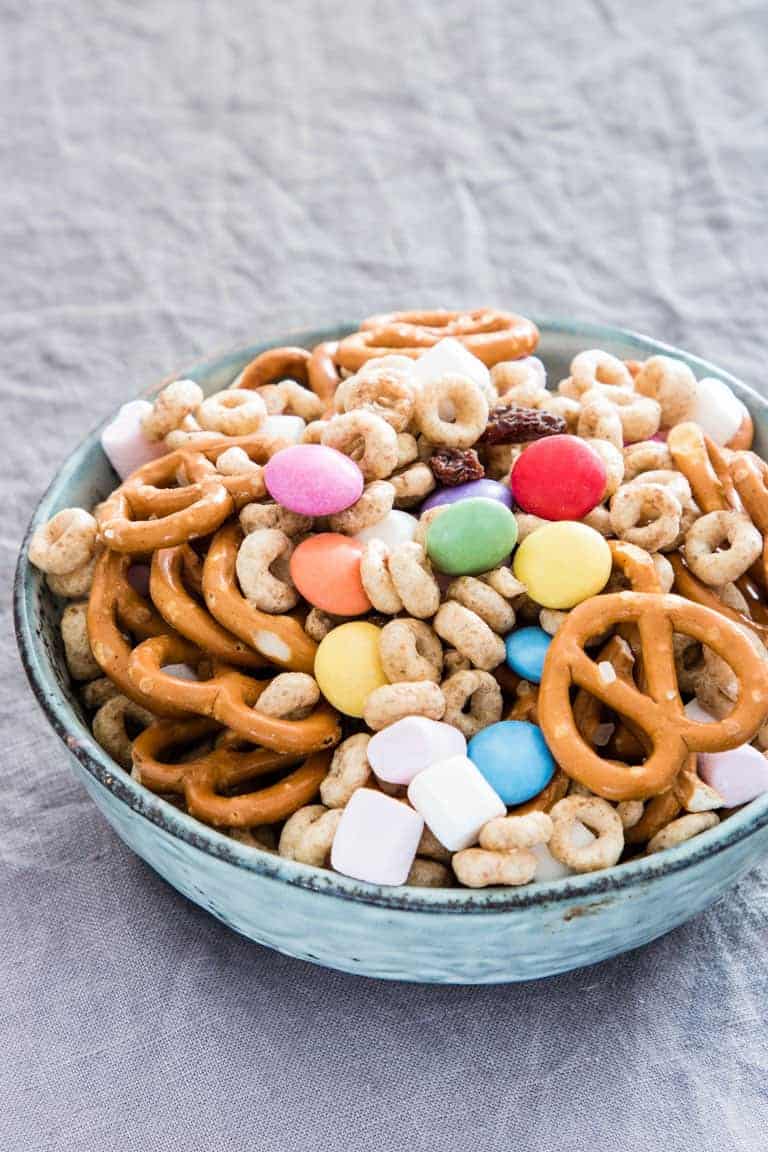 A bowl filled with pretzels, cereal rings, and colorful candies sits on a gray textured surface.