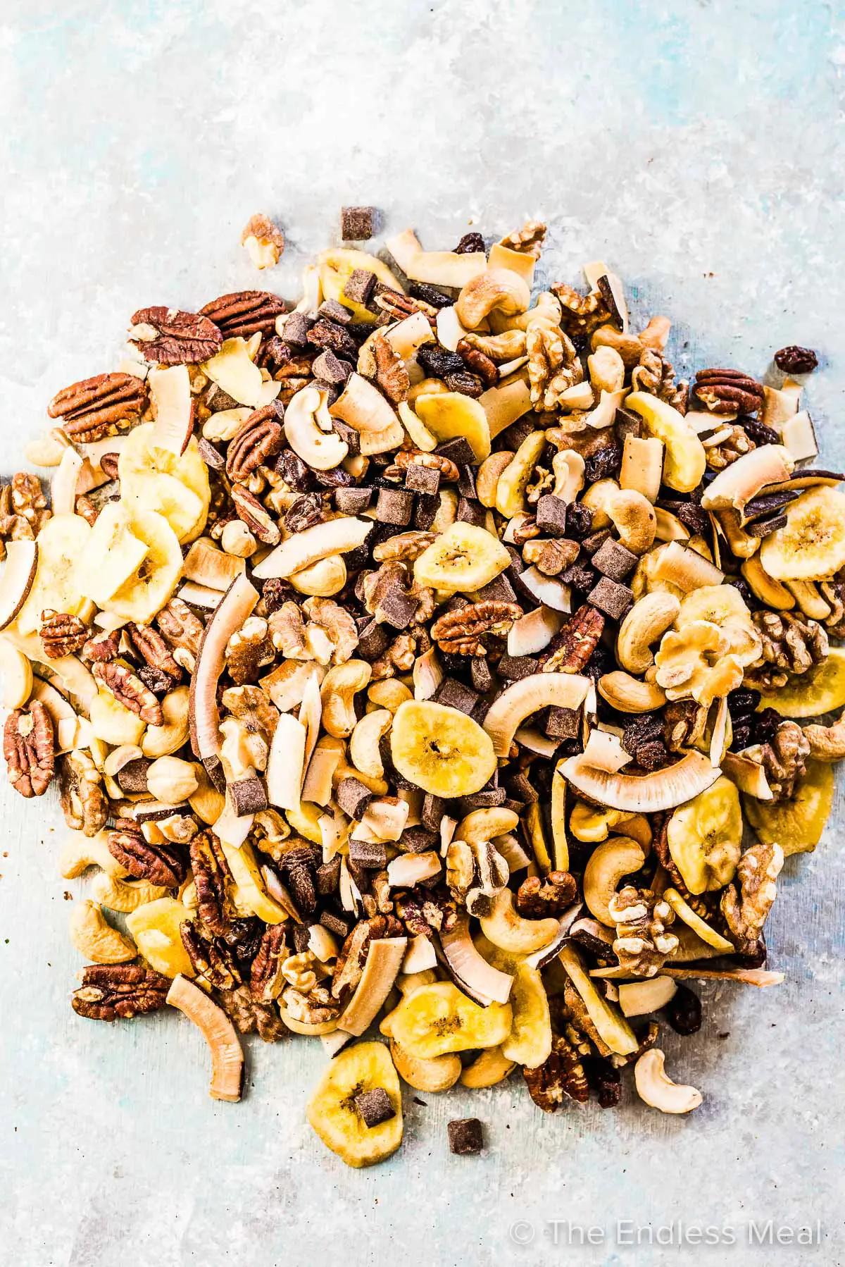 A colorful mix of chopped nuts, sliced bananas, coconut shavings, and chocolate chunks spread out on a light surface, creating a tempting composition indicative of a tasty granola or trail mix.