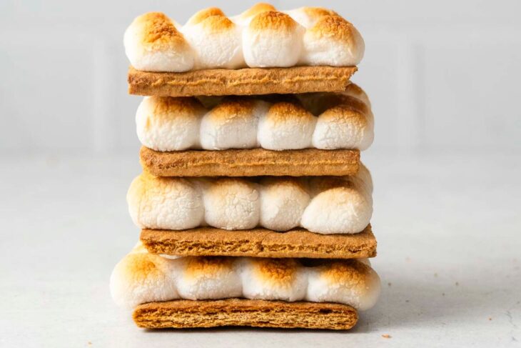 Four s'mores stacked with toasted marshmallows and graham crackers on a light-colored surface.