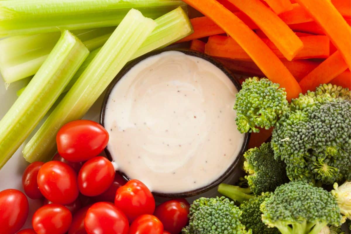 A bowl of dip surrounded by sliced carrots, celery, broccoli, and grape tomatoes.