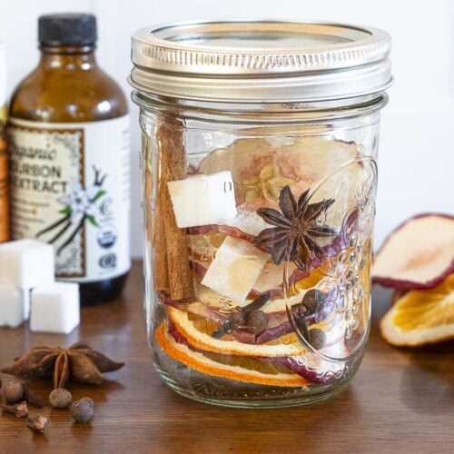A mason jar with dried orange and apple slices, star anise, cinnamon sticks, and spices sits on a table with bottles and sugar cubes.