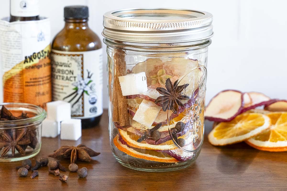 A mason jar with dried orange and apple slices, star anise, cinnamon sticks, and spices sits on a table with bottles and sugar cubes.
