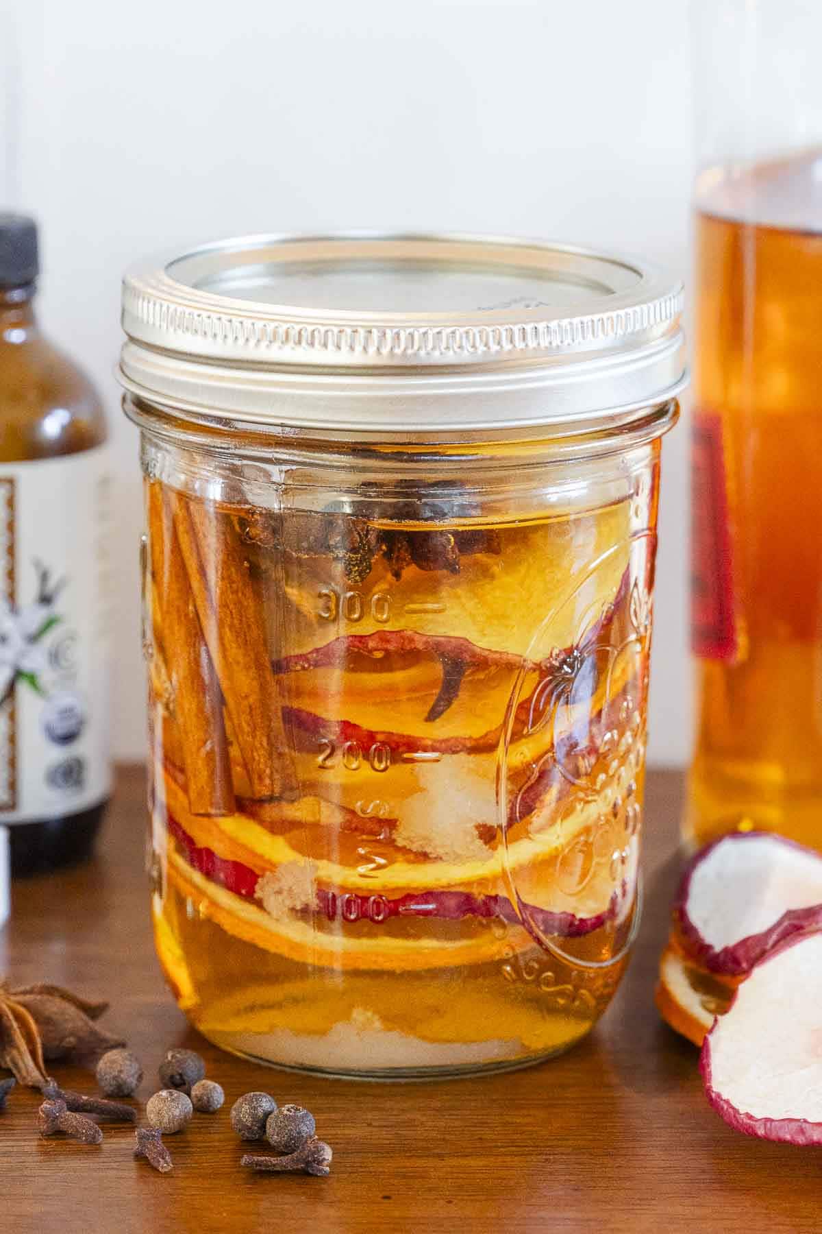 A mason jar with apple slices, cinnamon sticks, and spices in liquid sits on a wooden surface beside dried apples and spice bottles.