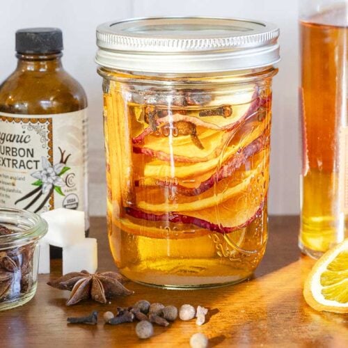 A mason jar with orange slices and spices on a wooden surface, surrounded by bottles, a bowl of star anise, sugar cubes, and an orange slice.