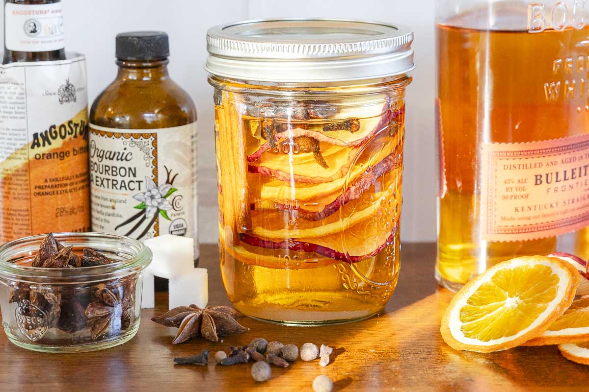 A mason jar with orange slices and spices on a wooden surface, surrounded by bottles, a bowl of star anise, sugar cubes, and an orange slice.