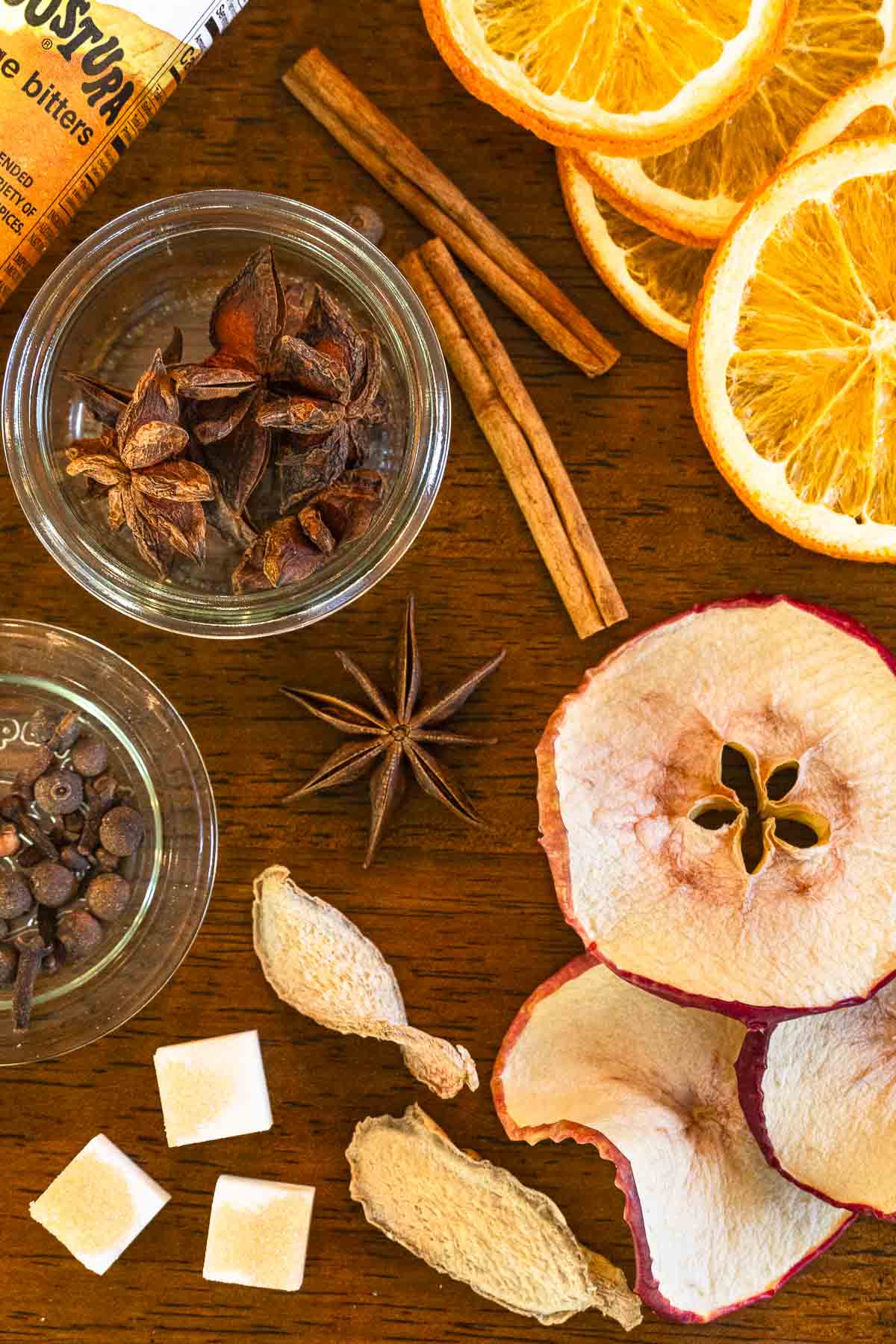 A flat lay of assorted dried fruit slices, spices, sugar cubes, and a bottle on a wooden surface.