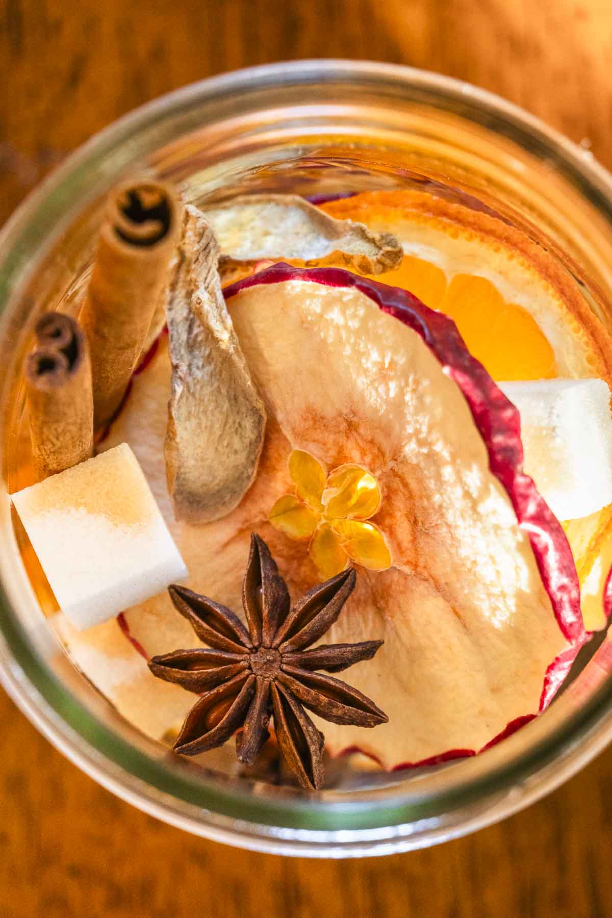 Top-down close-up of a jar filled with dried apple slices, star anise, cinnamon sticks, ginger, orange slices, and sugar cubes.