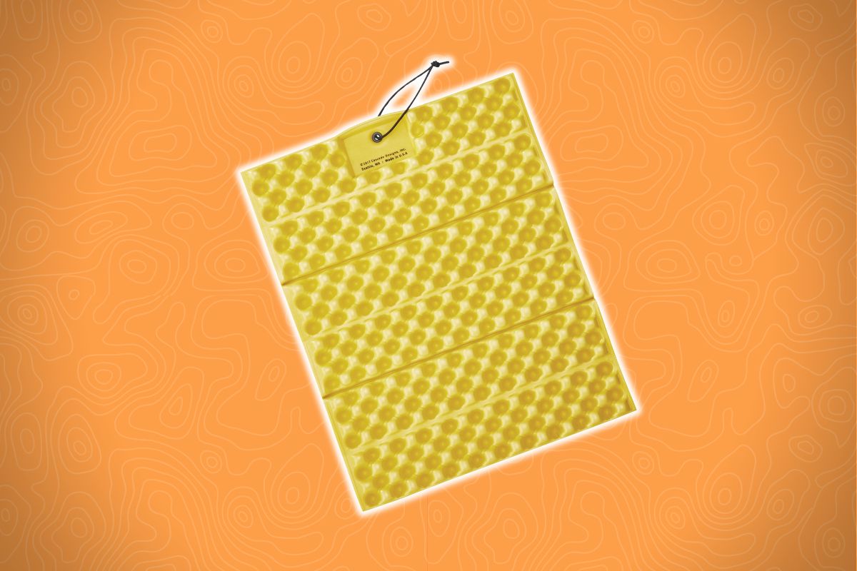 A yellow, foldable foam sleeping pad with a dimpled surface hangs by a black cord against an orange wavy-patterned background.