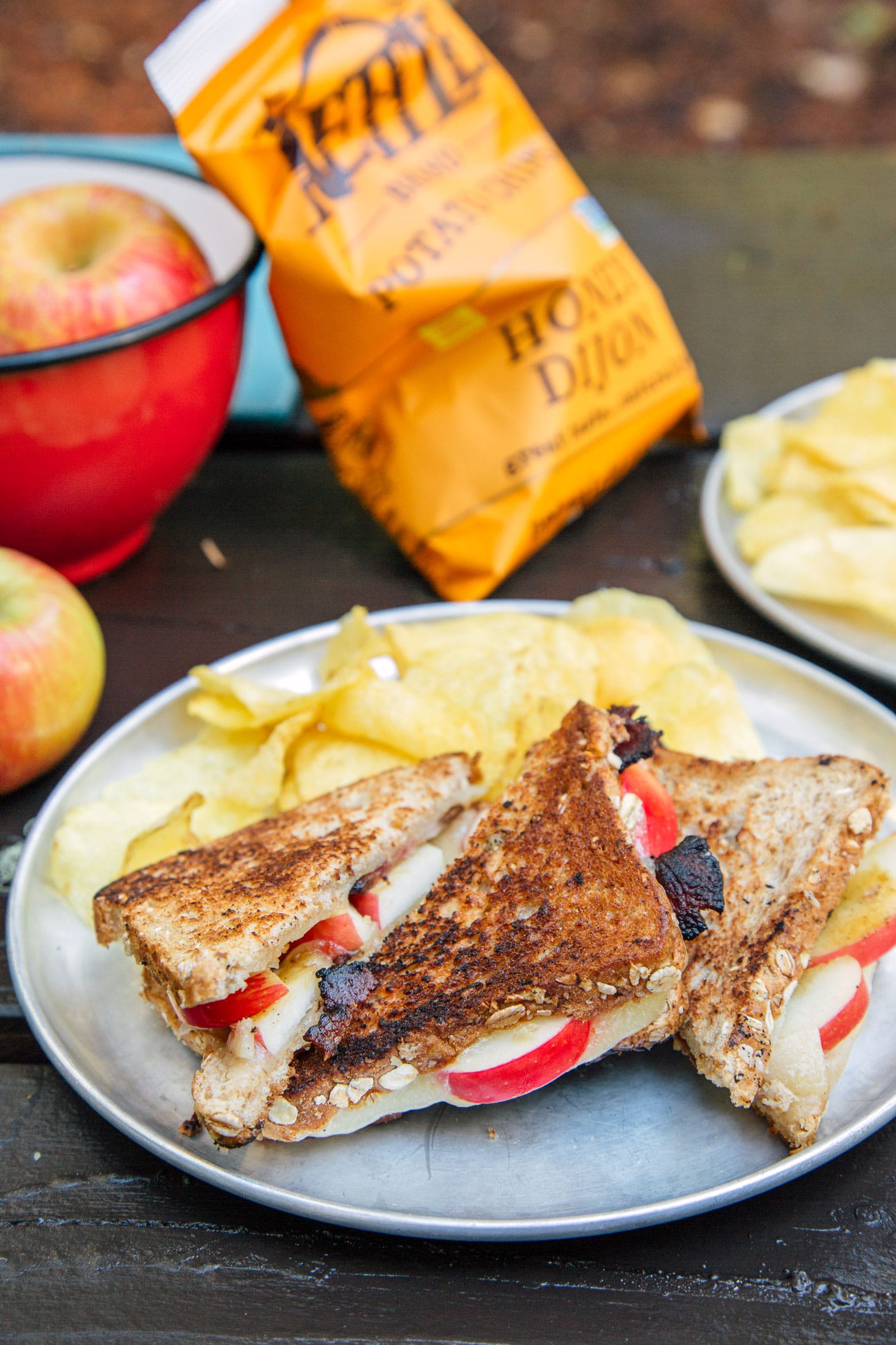A plate with grilled cheese and chips on a camp table. There is an orange chip bag and bowl of apples in the background.