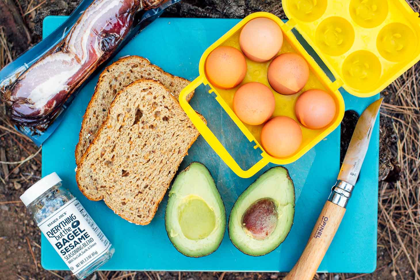 Ingredients to make an avocado toast breakfast sandwich (bacon, eggs, avocado, bread) on a blue cutting board