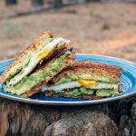 Breakfast sandwich with avocado, egg, and bacon on a blue camping plate placed on a log