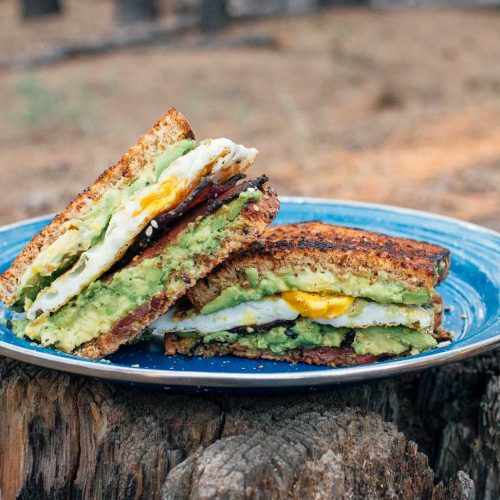 Breakfast sandwich with avocado, egg, and bacon on a blue camping plate placed on a log