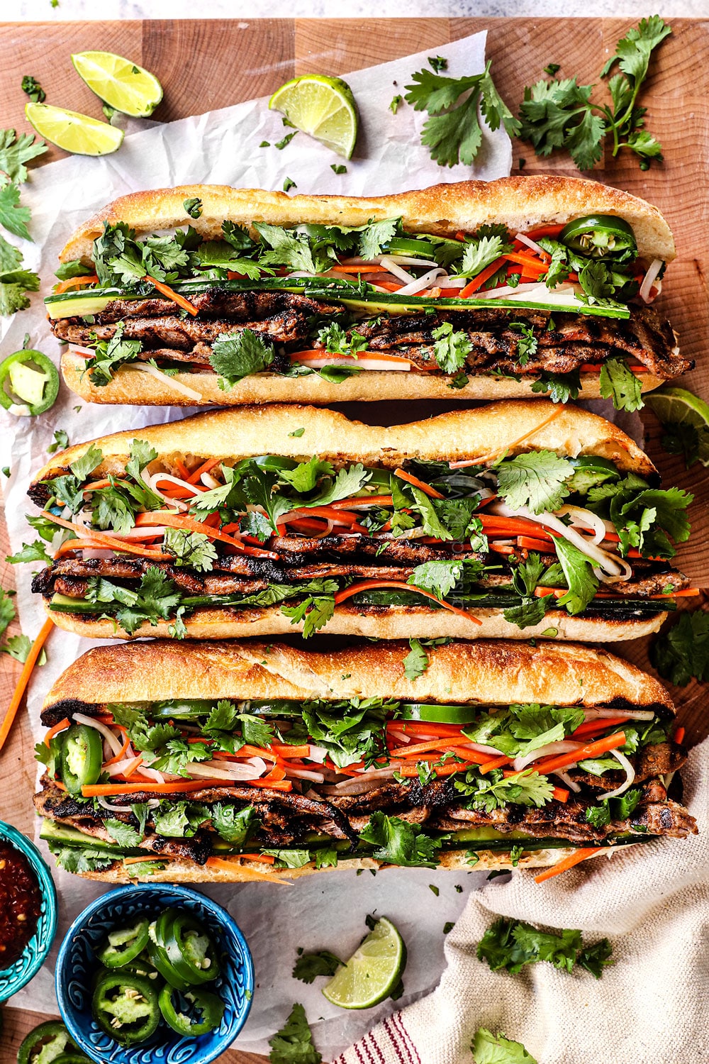 A banh mi sandwich is presented on a wooden table, showcasing a toasted baguette filled with grilled meats, pickled vegetables, fresh cilantro, jalapeño slices and accompanied by lime wedges.